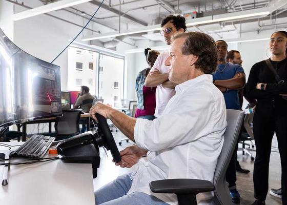 Jim Jones demonstrates a driving simulator with a steering wheel and two concave computer screens in the Cyber Lab at Mason Square